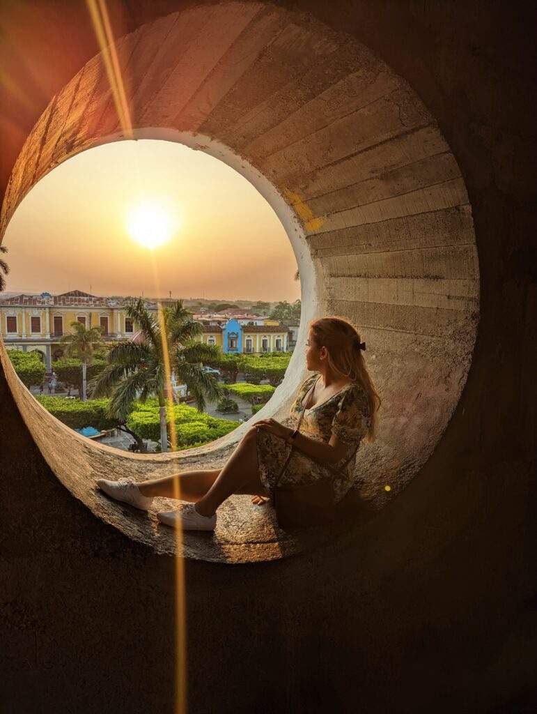 A woman in a dress sits on the circular window of a church tower looking down at Granada, Nicaragua during sunset.